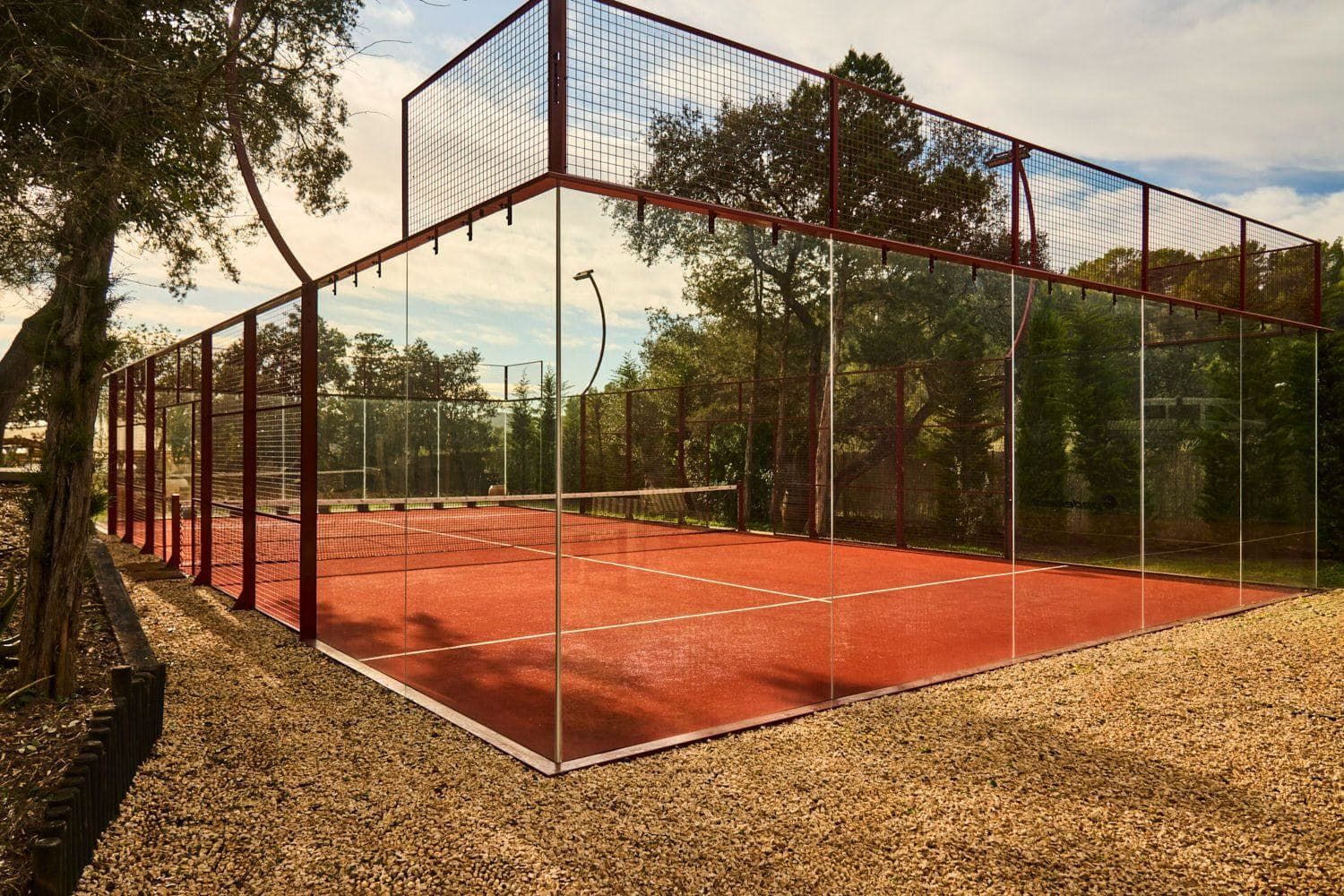 A padel court seen wide, clay-coloured surface, trees behind it