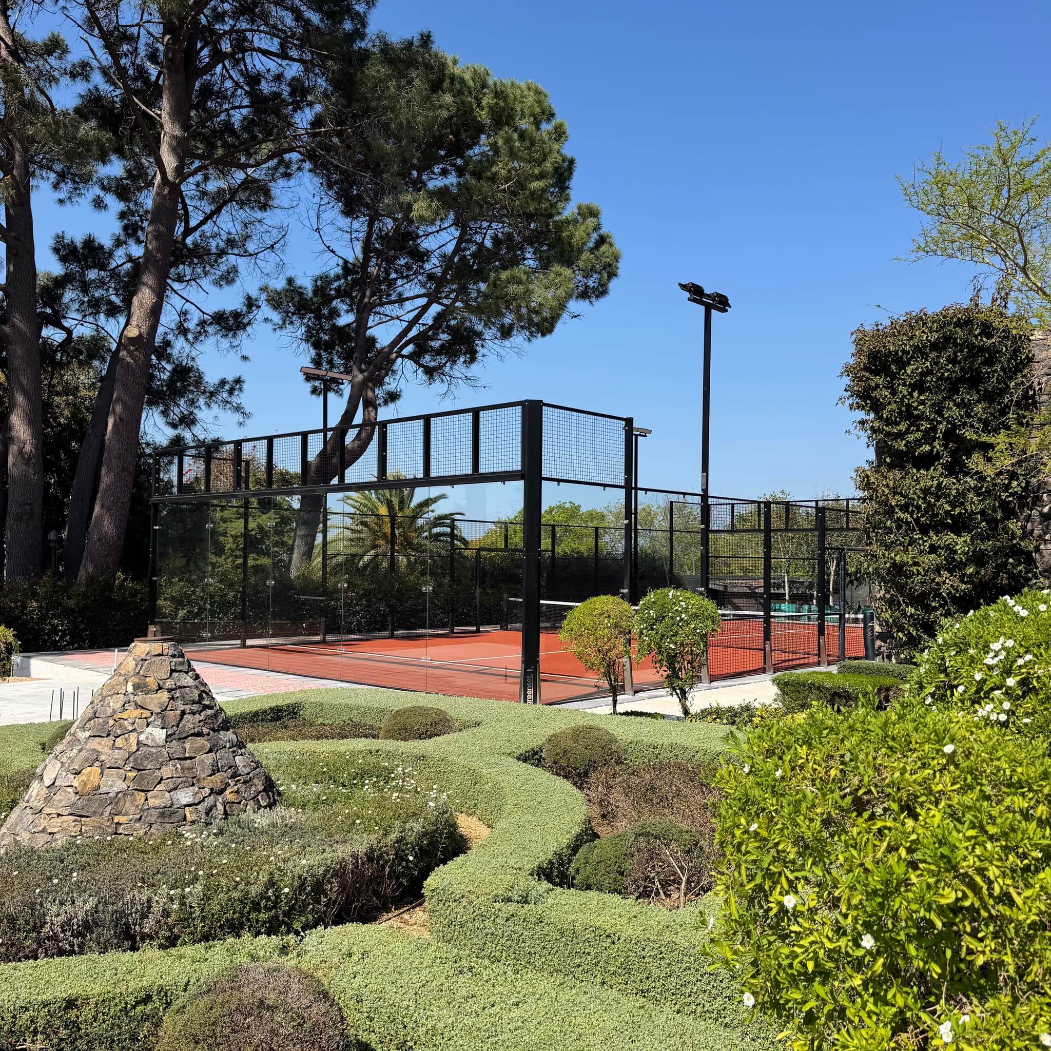 A padel court beyond a knot garden and its stone cairn